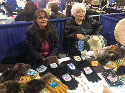 Two women vendors sitting at their booth, selling gloves that have decorative Native beading on them.